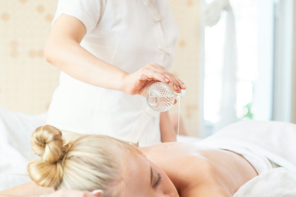 A woman lays on her stomach while an aromatherapy oil is poured on her back for a traditional massage